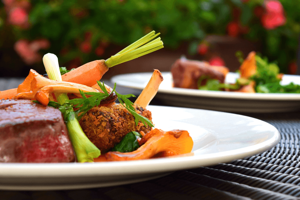 A gourmet plate featuring a slice of beef, sautéed vegetables, and a croquette, set against a blurred garden background.