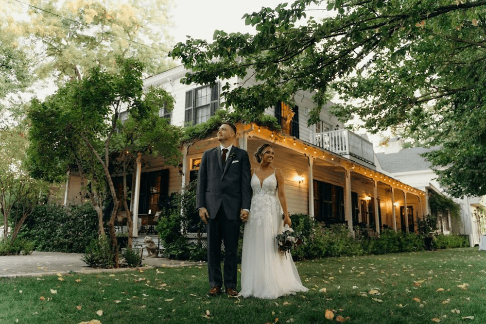 A newlywed couple stands hand-in-hand in front of a charming house adorned with warm lights.